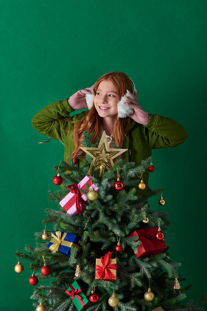 holidays, cheerful girl wearing ear muffs and standing behind decorated Christmas tree on turquoise - Fotografie, Obrázek
