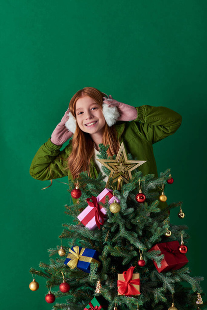 holiday spirit, happy girl wearing ear muffs and standing near decorated Christmas tree on turquoise - Photo, Image
