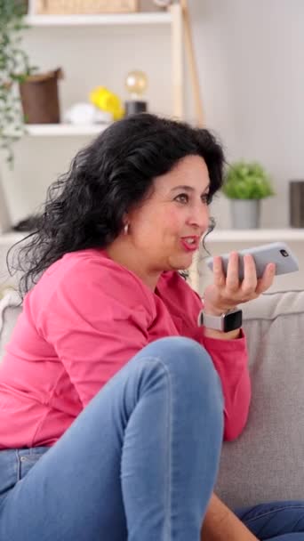 Mujer de mediana edad que envía un mensaje de voz en el teléfono mientras está sentado en el sofá - Imágenes, Vídeo