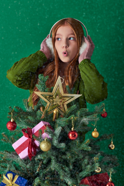 holiday spirit, surprised girl in ear muffs hugging decorated Christmas tree on turquoise backdrop - Photo, Image