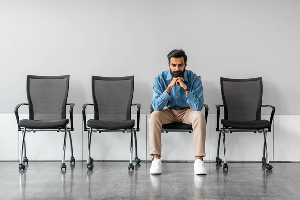 Долго ждал. Задумчивый серьезный индийский бизнесмен, держась за подбородок, сидя на стуле на фоне серой стены, полная длина, свободное место - Фото, изображение