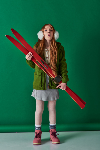 koude lucht, mooi meisje in oorkappen en winter outfit ademen en houden rode ski 's op turquoise - Foto, afbeelding