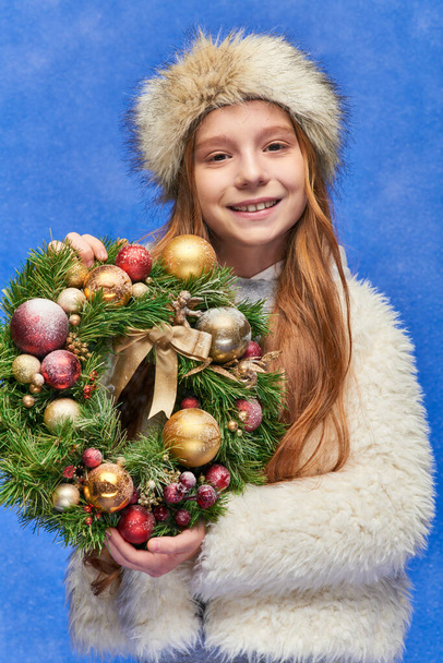 happy girl in faux fur hat and jacket holding Christmas wreath under falling snow on blue backdrop - Photo, Image
