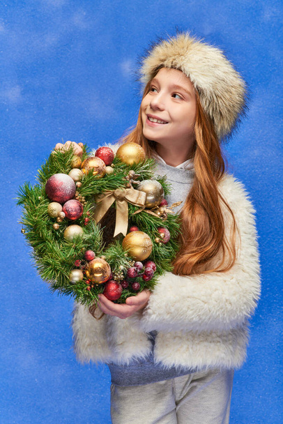 χαμογελαστό κορίτσι με ψεύτικο γούνινο καπέλο και σακάκι κρατώντας Χριστουγεννιάτικο στεφάνι κάτω από το χιόνι που πέφτει στο μπλε - Φωτογραφία, εικόνα
