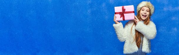 pancarta de invierno, chica alegre en chaqueta de piel sintética y sombrero celebración caja de regalo sobre fondo turquesa - Foto, Imagen