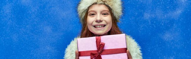winter holiday banner, happy girl in faux fur jacket and hat holding wrapped gift box on turquoise - Photo, Image