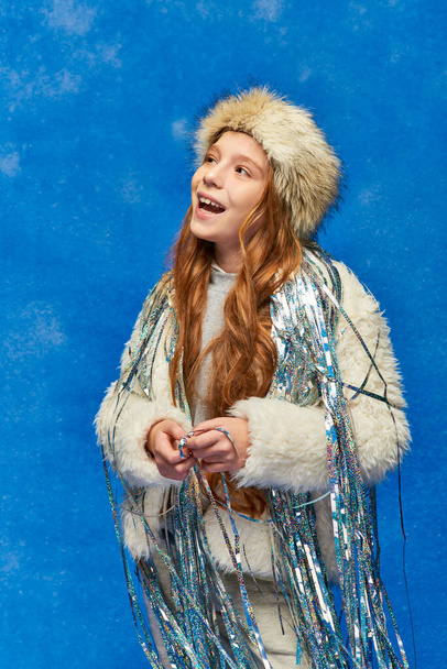 allegra ragazza in pelliccia sintetica giacca e cappello con fili di lame in piedi sotto la neve caduta su sfondo blu - Foto, immagini