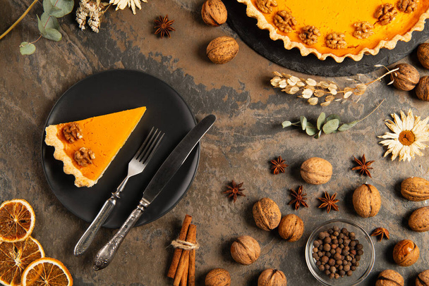 maukas kurpitsapiirakka ja ruokailuvälineet mustalla keraamisella lautasella lähellä oranssia kurpitsaa kivipöydällä, kiitospäivä - Valokuva, kuva