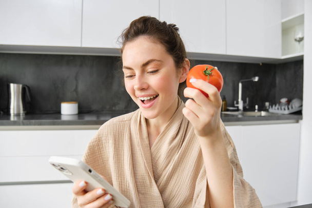 Close up portrait of beautiful young woman in the kitchen, holding tomato and smartphone, buys fresh vegetables online, orders groceries delivery on mobile phone application. - Photo, Image