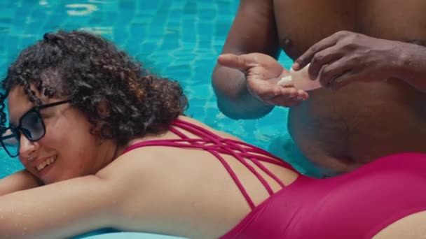 Средний снимок счастливой улыбающейся молодой белой девушки, отдыхающей в бассейне на надувном матрасе, и афро-американского парня, распространяющего солнцезащитный крем на ее спине - Кадры, видео