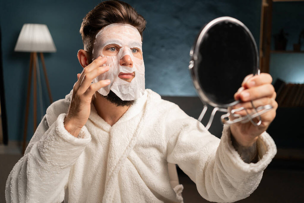 Hombre barbudo hispano con máscara de sábanas en la cara, haciendo tratamiento de belleza en casa y mirando al espejo. Rutina de belleza, concepto de autocuidado. - Foto, Imagen