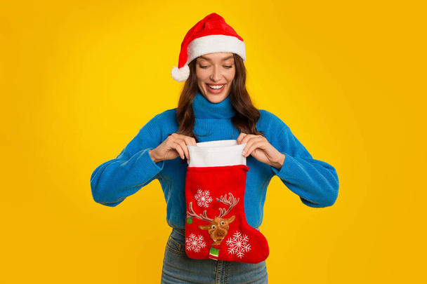 Xmas Surpresas. Happy European Lady In Santa Hat Looking into Christmas Stocking Sock Over Yellow Studio Background (em inglês). Férias de Inverno Temporada Celebração e ofertas Conceito - Foto, Imagem