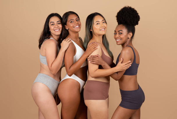 Vier verschillende jonge dames die vol vertrouwen poseren in hun ondergoed, natuurlijke schoonheid en eenheid vieren op beige studio-achtergrond, kijkend en glimlachend naar de camera - Foto, afbeelding