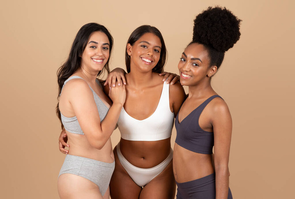 Três mulheres multirraciais posando confortavelmente em suas cuecas, retratando momento genuíno e íntimo no estúdio, fundo bege, senhoras comemorando a beleza autêntica e amizade - Foto, Imagem