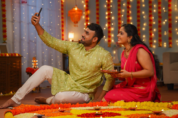 Casais indianos felizes tirando selfie no telefone celular durante a celebração do festival diwali na frente de rangoli em casa conceito de decoração de flores, compartilhamento de mídia social e relacionamento. - Foto, Imagem