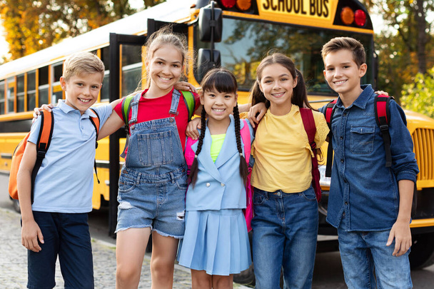 Счастливые улыбающиеся дети, ожидающие рядом с желтым школьным автобусом, группа веселых детей в рюкзаках, готовых идти домой после дня учебы и веселья, позитивные ученики, улыбающиеся и глядящие в камеру - Фото, изображение