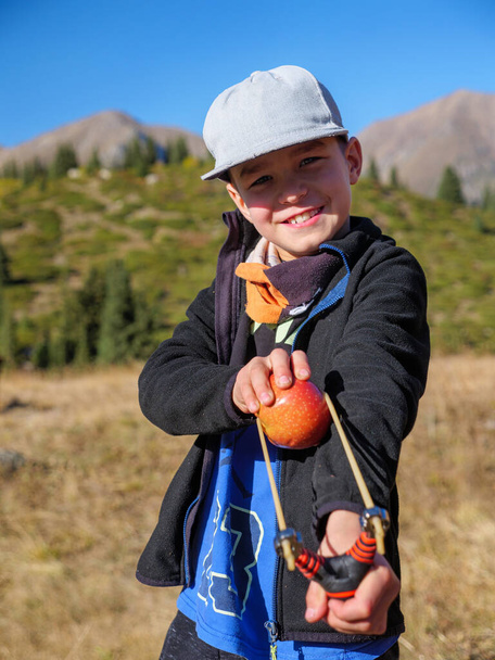 Junge will einen großen roten Apfel aus einer Schleuder schießen. Verdorben junge having spaß draußen. - Foto, Bild