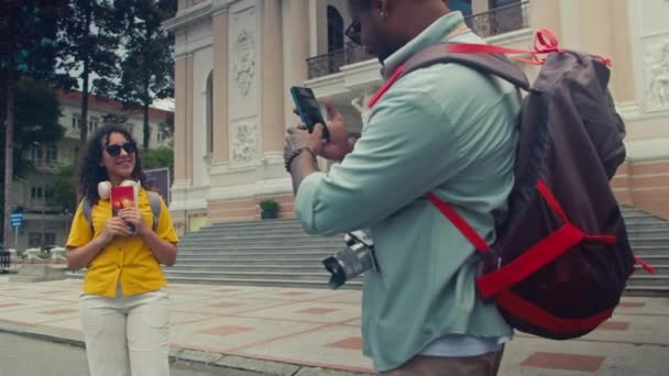 Mittlere Gesamtaufnahme einer jungen kaukasischen Frau, die vor dem Opernhaus in Ho-Chi-Minh-Stadt in Vietnam steht, ein Touristenheft in der Hand hält und mit ihrem schwarzen Freund ein Video auf dem Smartphone für den Reiseblog aufzeichnet - Filmmaterial, Video
