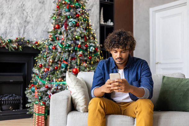 Poważne myślenie człowiek koncentruje się na wyborze prezentów i towarów w sklepie internetowym, latynoski siedzi w domu na kanapie na Boże Narodzenie, trzymając smartfona, w pobliżu drzewa na Nowy Rok wakacje. - Zdjęcie, obraz