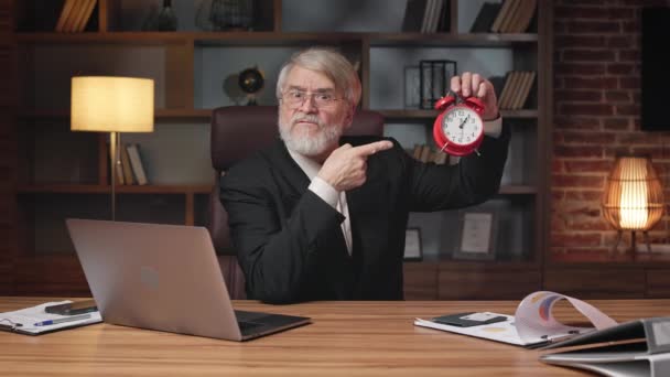 Resentful older male in formal clothes demonstrating mechanical alarm clock while talking to camera in workplace. Irritated business person appealing to tight deadlines of construction project. - Footage, Video