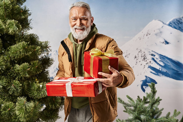 happy Santa in warm jacket holding presents near pine tree with mountains backdrop, winter concept - Photo, Image
