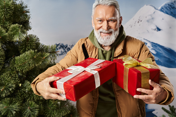 przystojny Mikołaj z białą brodą w ciepłej kurtce trzyma prezenty i uśmiecha się wesoło, zima - Zdjęcie, obraz