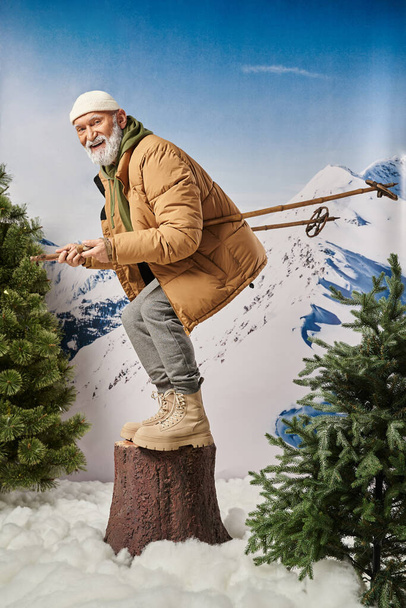 Père Noël avec barbe blanche accroupi sur tronc d'arbre avec bâtons de ski souriant à la caméra, concept d'hiver - Photo, image