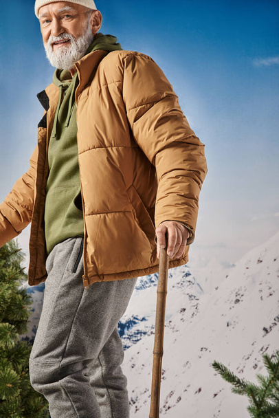 guapo Santa con barba blanca mirando alegremente a la cámara posando con bastones de esquí, concepto de invierno - Foto, imagen