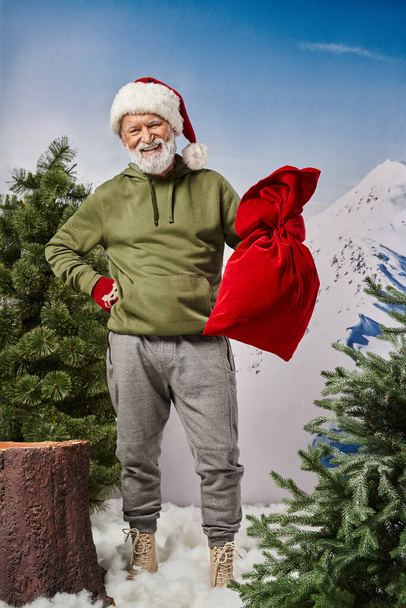 vidám férfi khaki kapucnis pulcsiban, kezében Mikulás táska és visel karácsonyi kalap, Boldog Karácsonyt - Fotó, kép