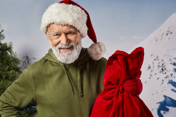 homem atlético alegre com capuz cáqui vestindo chapéu de Papai Noel e segurando saco presente, conceito de inverno - Foto, Imagem