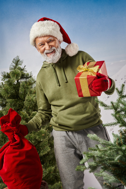 joyful Santa holding present bag and red gift in hands and smiling at camera, winter concept - Foto, Imagem
