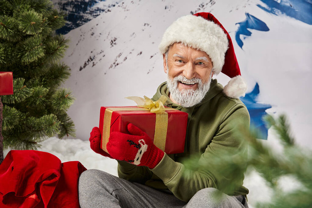 athletic Santa in red hat and mittens sitting with gift on snow near present bag, winter concept - Photo, Image