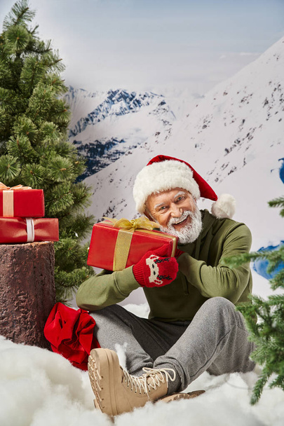 cheerful man dressed as Santa holding present and sitting on snow near tree stump, winter concept - Photo, Image