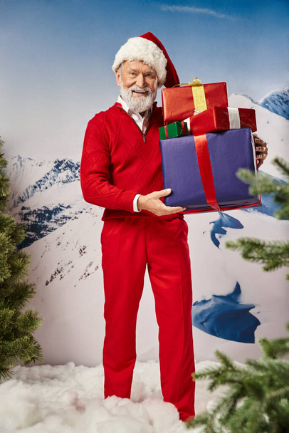 cheerful man in red Santa costume with presents in hands with snowy backdrop, winter concept - Fotó, kép