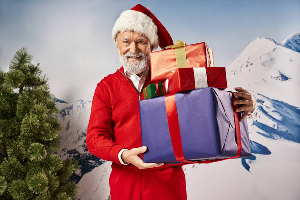 fehér szakállas Mikulás gazdaság ajándékok és mosolygós kamera havas háttér, téli koncepció - Fotó, kép