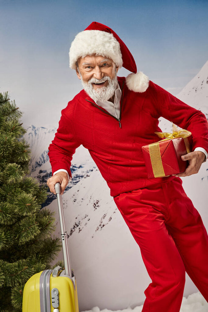 χαρούμενο κοστούμι Santa με λευκή γενειάδα ποζάρουν με κίτρινη βαλίτσα και παρουσιάζουν, Χριστούγεννα έννοια - Φωτογραφία, εικόνα