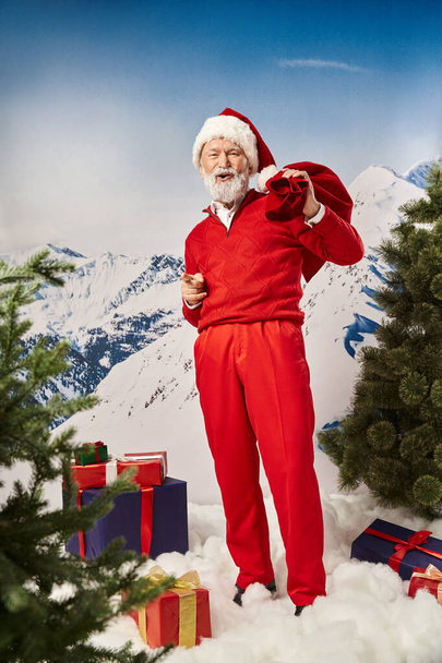 gelukkig kerstman met geschenk zak gebaar omgeven door geschenken met open mond, winter concept - Foto, afbeelding