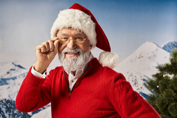 homem alegre vestido como Papai Noel em chapéu vermelho tocando seus óculos e olhando para a câmera, Feliz Natal - Foto, Imagem