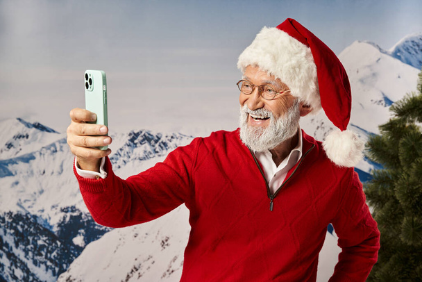 homem feliz vestido de Papai Noel vestindo óculos e tomando selfie com pano de fundo de montanha, conceito de inverno - Foto, Imagem