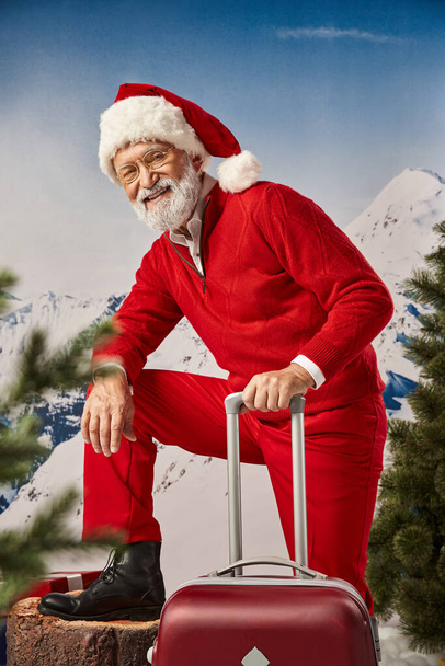 χαρούμενος άνθρωπος ντυμένος Άγιος Βασίλης σε γυαλιά που ποζάρουν με βαλίτσα κοιτάζοντας την κάμερα, έννοια του χειμώνα - Φωτογραφία, εικόνα