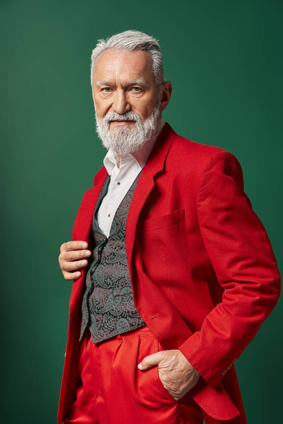 retrato de Papá Noel elegante en traje rojo elegante posando con una mano en el bolsillo, concepto de invierno - Foto, imagen