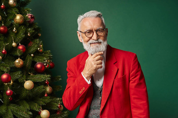 jolly Santa in glasses with beard in classy suit posing near tree with hand on chin, winter concept - 写真・画像