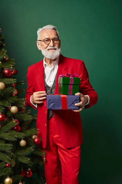 elegancki Mikołaj w stylowym garniturze z prezentami i patrząc na kamerę na zielonym tle, koncepcja zima - Zdjęcie, obraz
