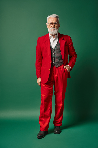 classy man dressed as Santa posing with one hand in pocket on green backdrop, winter concept - Photo, Image