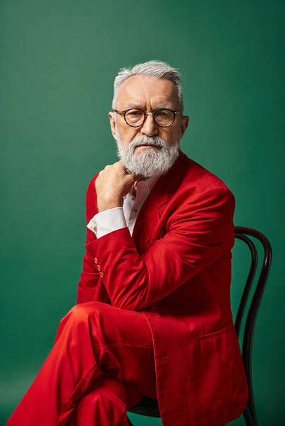 élégant beau Père Noël en costume rouge posant sur la chaise avec sa main près du visage, concept de Noël - Photo, image