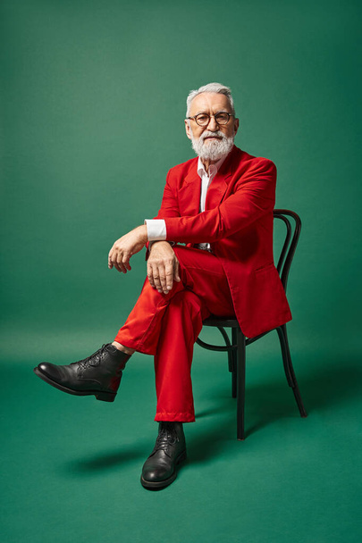 classy good looking Santa in red suit sitting on chair on dark green backdrop, winter concept - Фото, зображення
