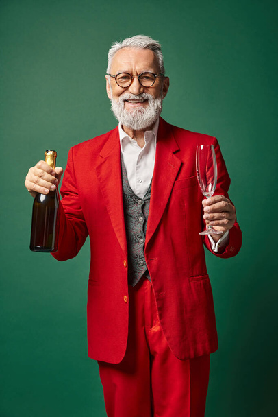 Papai Noel feliz com óculos e barba em terno vermelho posando com garrafa de champanhe, conceito de inverno - Foto, Imagem