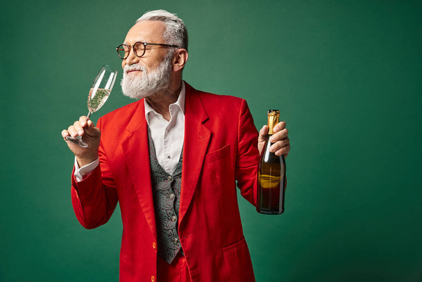 elegant Santa with beard in red classy suit testing champagne on green backdrop, winter concept - Photo, Image
