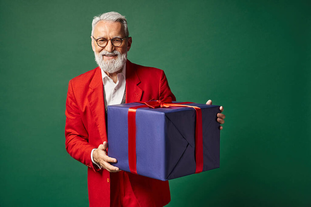 элегантный Санта с бородой и очками держа огромный подарок и улыбаясь искренне, зимняя концепция - Фото, изображение