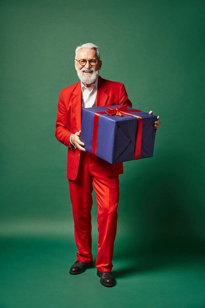 gut aussehender Mann in Weihnachtsmannkleidung posiert mit riesigem Geschenk auf grünem Hintergrund, Winterkonzept - Foto, Bild